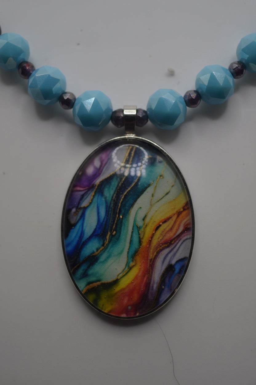 A beaded necklace with a colorful oval pendant, displayed on a light gray surface. The necklace features a silver chain, white seed beads, light blue beads, and small accent beads in green, orange, and black, culminating in a marbled multicolor pendant.