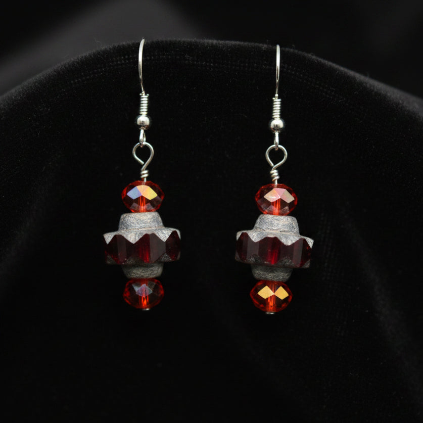 A pair of handmade dangle earrings with faceted dark gray beads, orange-red crystal beads, and silver-tone hooks, displayed on a clear stand against a neutral gray background.
