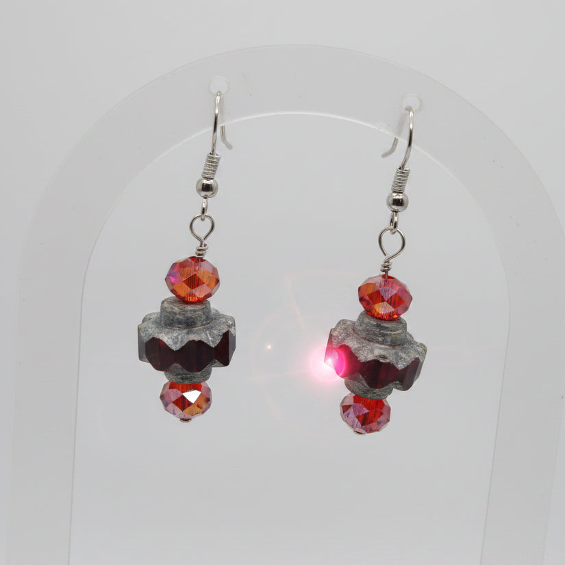 A pair of handmade dangle earrings featuring textured dark gray beads, small red faceted beads, and silver fishhook wires, displayed on a clear plastic stand against a neutral gray background.