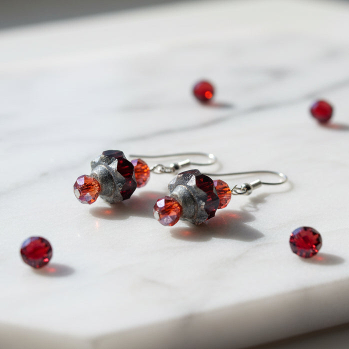 A pair of handmade dangle earrings with faceted red beads, textured dark gray stone beads, and silver fishhook wires, displayed on a clear plastic stand against a neutral gray background.