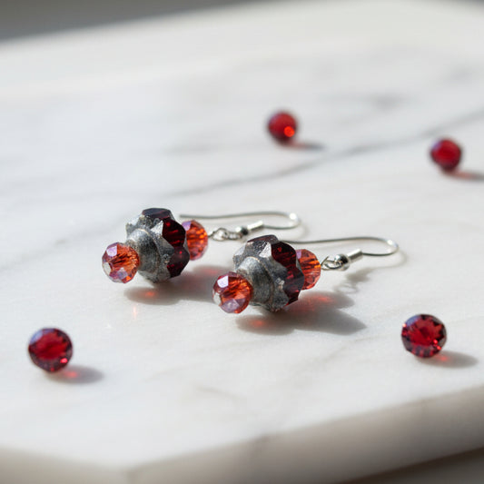 A pair of handmade dangle earrings with faceted red beads, textured dark gray stone beads, and silver fishhook wires, displayed on a clear plastic stand against a neutral gray background.