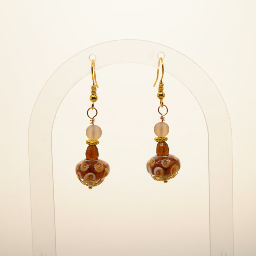 A pair of gold-tone dangle earrings with patterned amber and white glass beads, displayed on a clear acrylic stand against a neutral gray background.