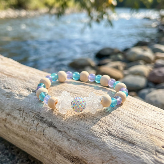 A beaded stretch bracelet featuring light blue and white beads, natural wood beads, and a central clear crystal charm, displayed on a neutral gray background.