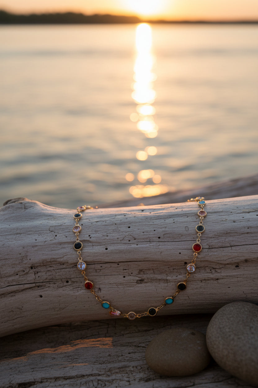 A gold-toned link necklace featuring multiple small round gemstones in assorted colors including blue, red, black, and clear, displayed on a neutral gray background.