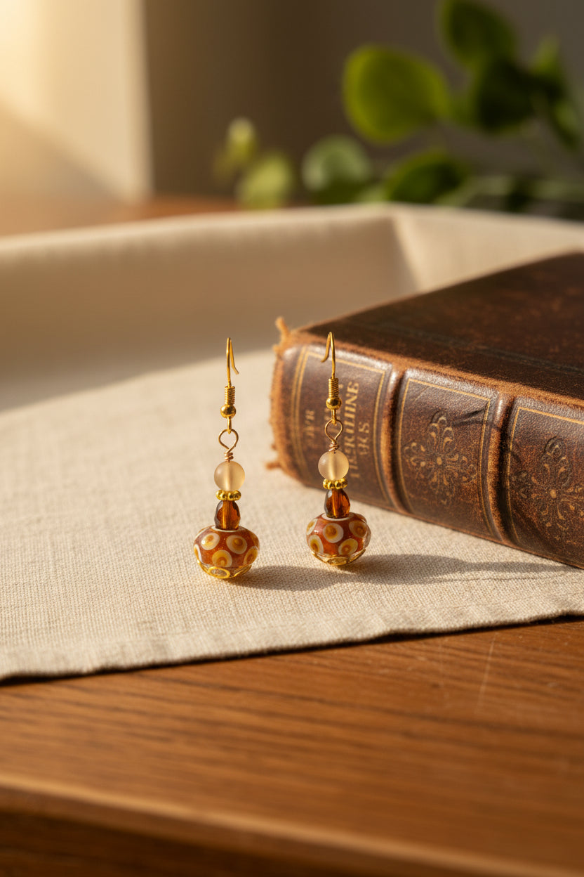 A pair of handmade glass drop earrings with gold-tone ear wires, a small pale pink bead, a translucent brown bead, and a larger patterned brown glass bead at the bottom, displayed on a clear acrylic stand against a light gray background.