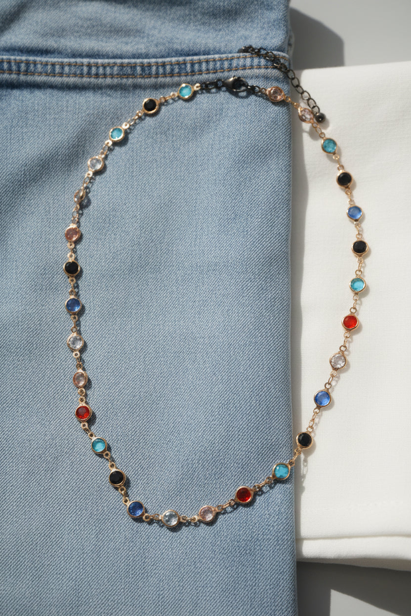 A delicate gold-toned necklace featuring small, alternating round gemstones in red, light blue, dark blue, and black, arranged in a loose oval on a neutral gray background.