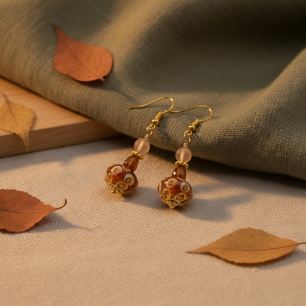 A pair of handmade glass lampwork dangle earrings with gold-toned hooks, clear beads, and brown patterned glass pendants, displayed on a clear acrylic stand against a neutral gray background.