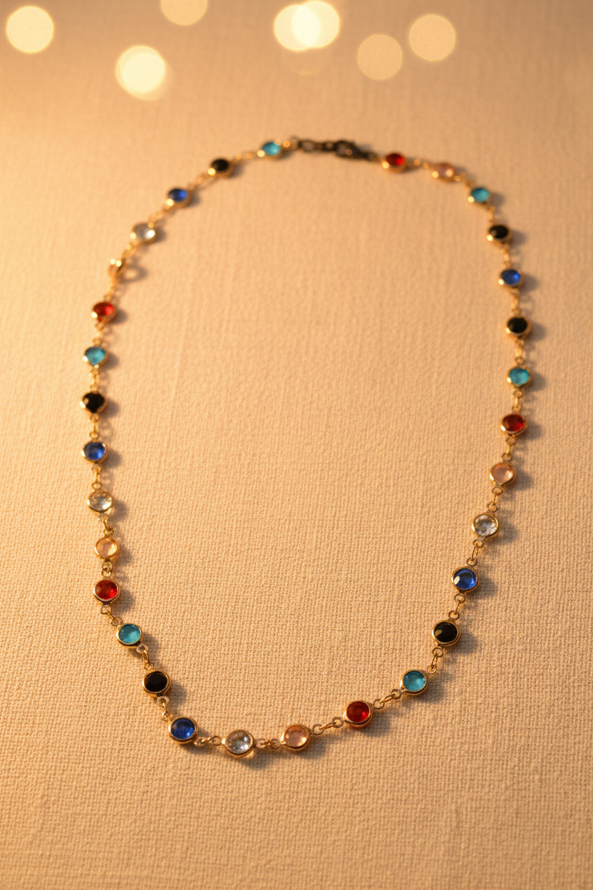 A delicate gold chain necklace featuring small, colorful round bezel-set gemstones in various colors including blue, red, black, and light blue, displayed on a neutral gray background.
