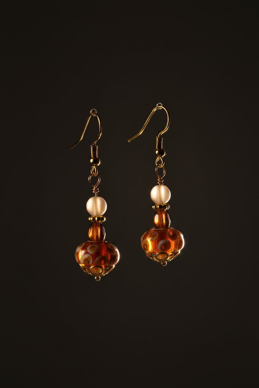 A pair of handmade glass lampwork dangle earrings with gold fishhook wires, clear round beads, and patterned brown glass drops on a neutral gray background.