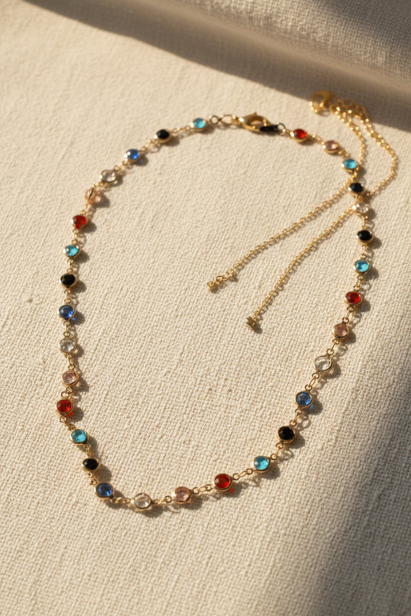 A gold-toned beaded necklace with small round gemstone accents in red, blue, black, and clear, arranged in a circle on a neutral gray background.