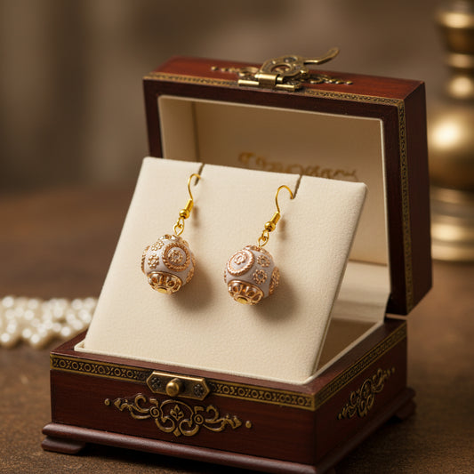 A pair of dangling earrings with gold-toned hooks and spherical filigree charms in a warm metallic rose-gold/copper tone, displayed on a clear stand against a soft gray background.