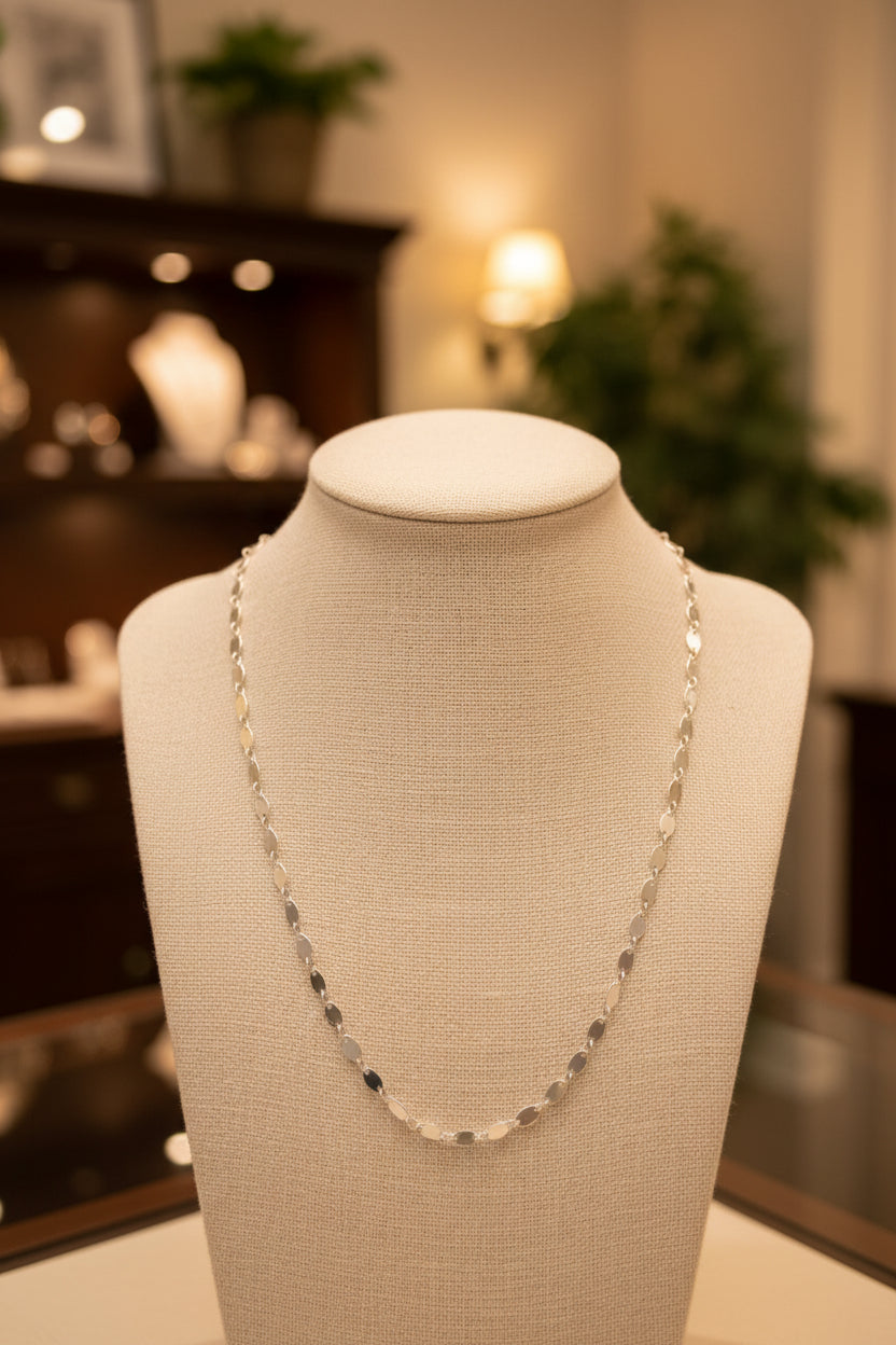 A silver chain necklace with small, flat, oval-shaped links, displayed against a plain light gray background.