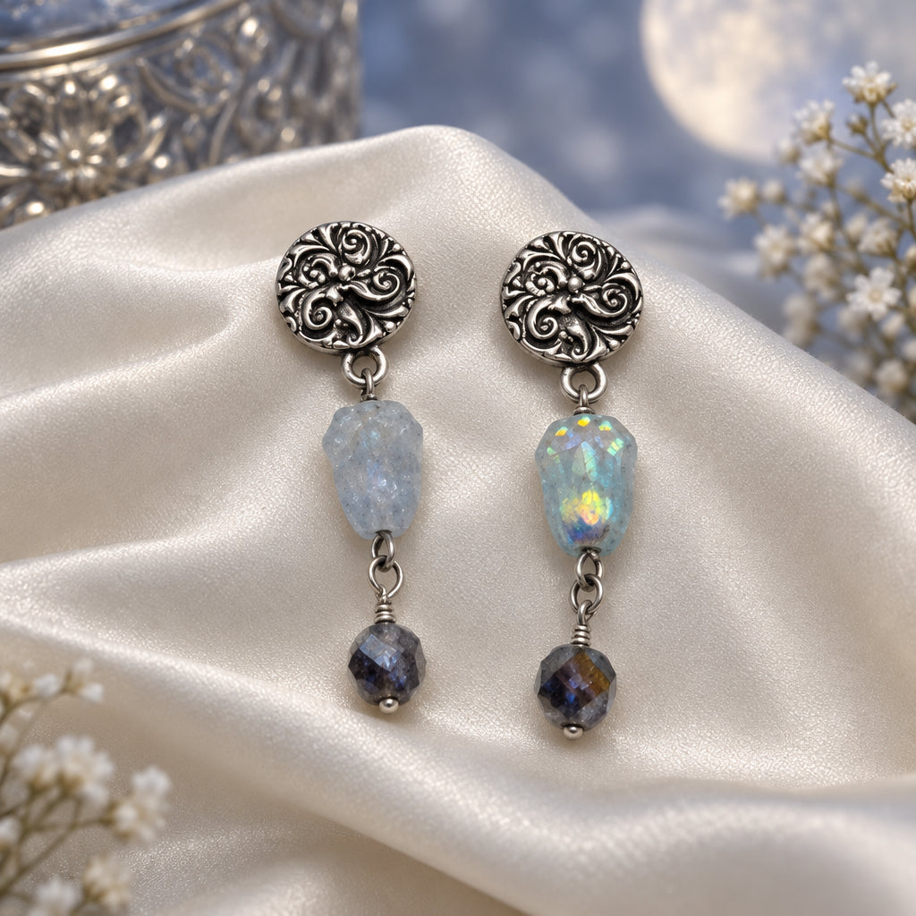A pair of dangling earrings on a white display card. The earrings have ornate silver-toned filigree tops, light blue faceted beads, iridescent beads, and dark brown beads at the bottom.