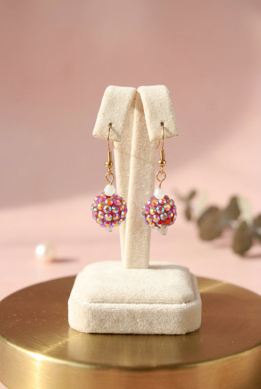 A pair of gold-tone dangle earrings with iridescent, rainbow-reflective rhinestone-covered round drops, displayed on a clear acrylic stand against a light gray background.