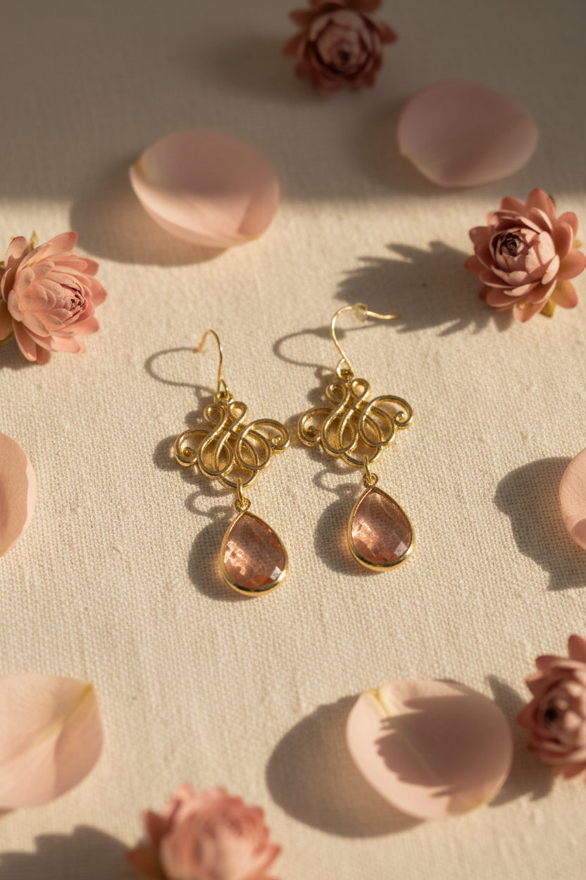A pair of gold-tone filigree dangle earrings with pink teardrop gemstones, displayed on a white 'River Inspired' jewelry card against a plain light gray background.