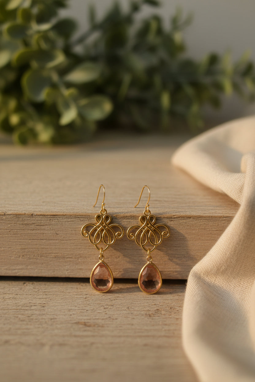 A pair of gold-tone drop earrings with ornate filigree tops and teardrop-shaped pink stones, displayed on a white card labeled River Inspired.