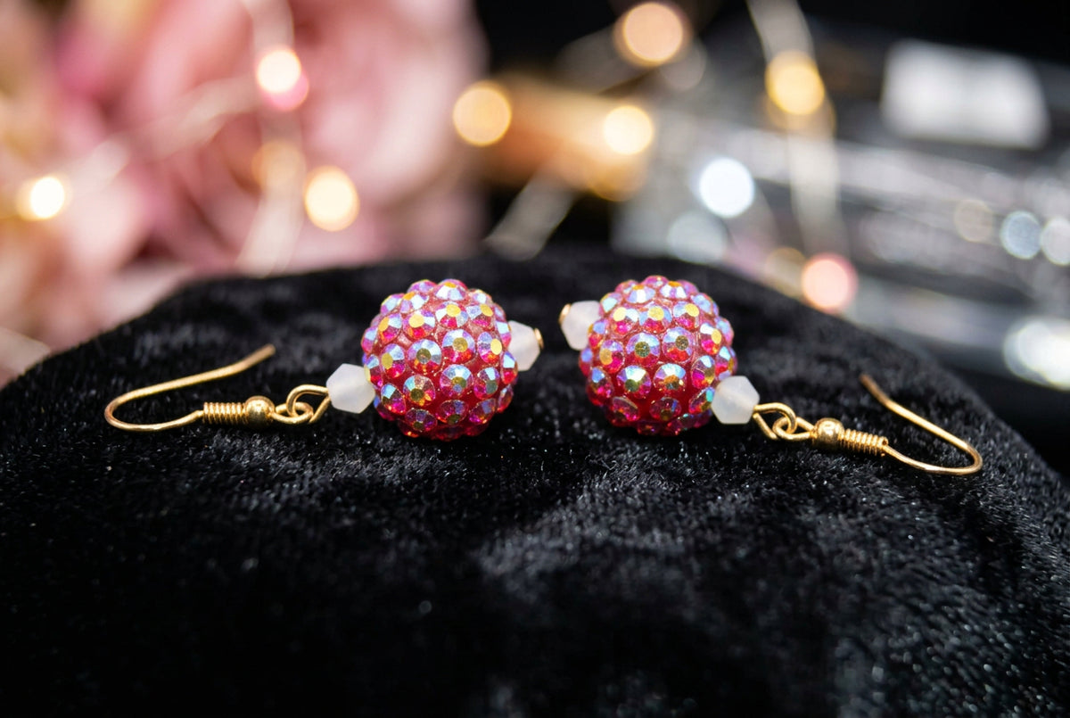 A pair of gold-tone dangle earrings with heart-shaped charms covered in iridescent, rainbow-reflecting rhinestones, displayed on a clear plastic stand against a light gray background.