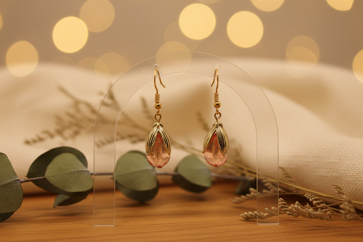 A pair of dangling earrings with gold-toned hooks and textured gold-toned leaf-like drops, each holding a translucent pink teardrop-shaped stone, displayed against a light gray background.