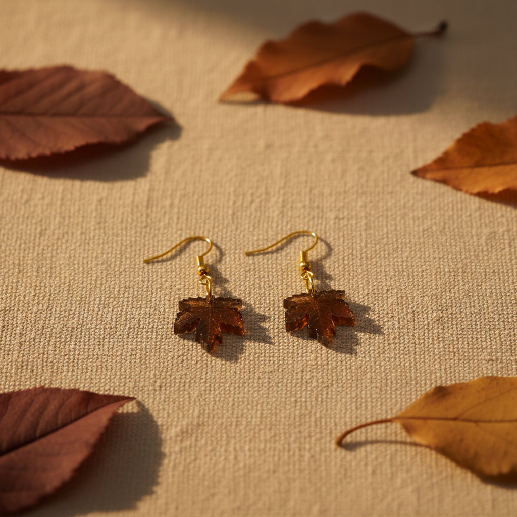 A pair of dangling earrings with small gold fishhook wires and leaf-shaped charms in a warm brown tone, displayed against a plain light gray background.