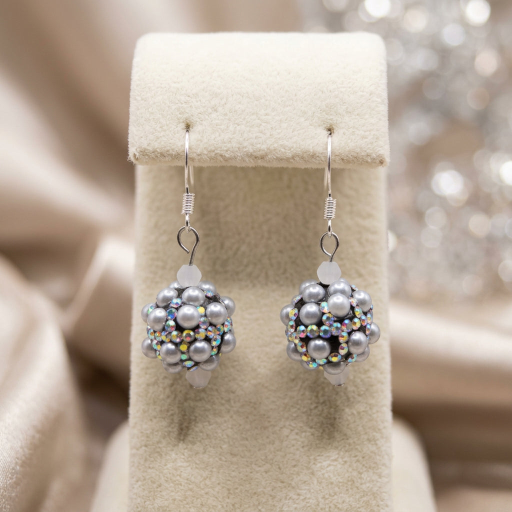 A pair of handmade beaded dangle earrings with gray faux pearls and iridescent beads, hanging from silver fishhook wires on a clear plastic display stand against a neutral gray background.