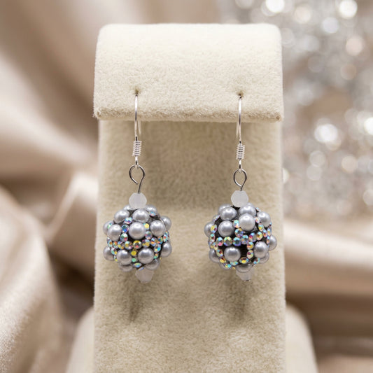 A pair of handmade beaded dangle earrings with gray faux pearls and iridescent beads, hanging from silver fishhook wires on a clear plastic display stand against a neutral gray background.