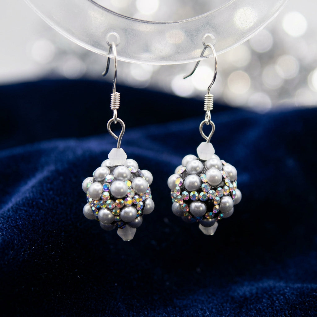 A pair of handmade beaded dangle earrings with silver fishhook wires, featuring gray faux pearls and iridescent AB beads, displayed on a clear acrylic stand against a neutral gray background.