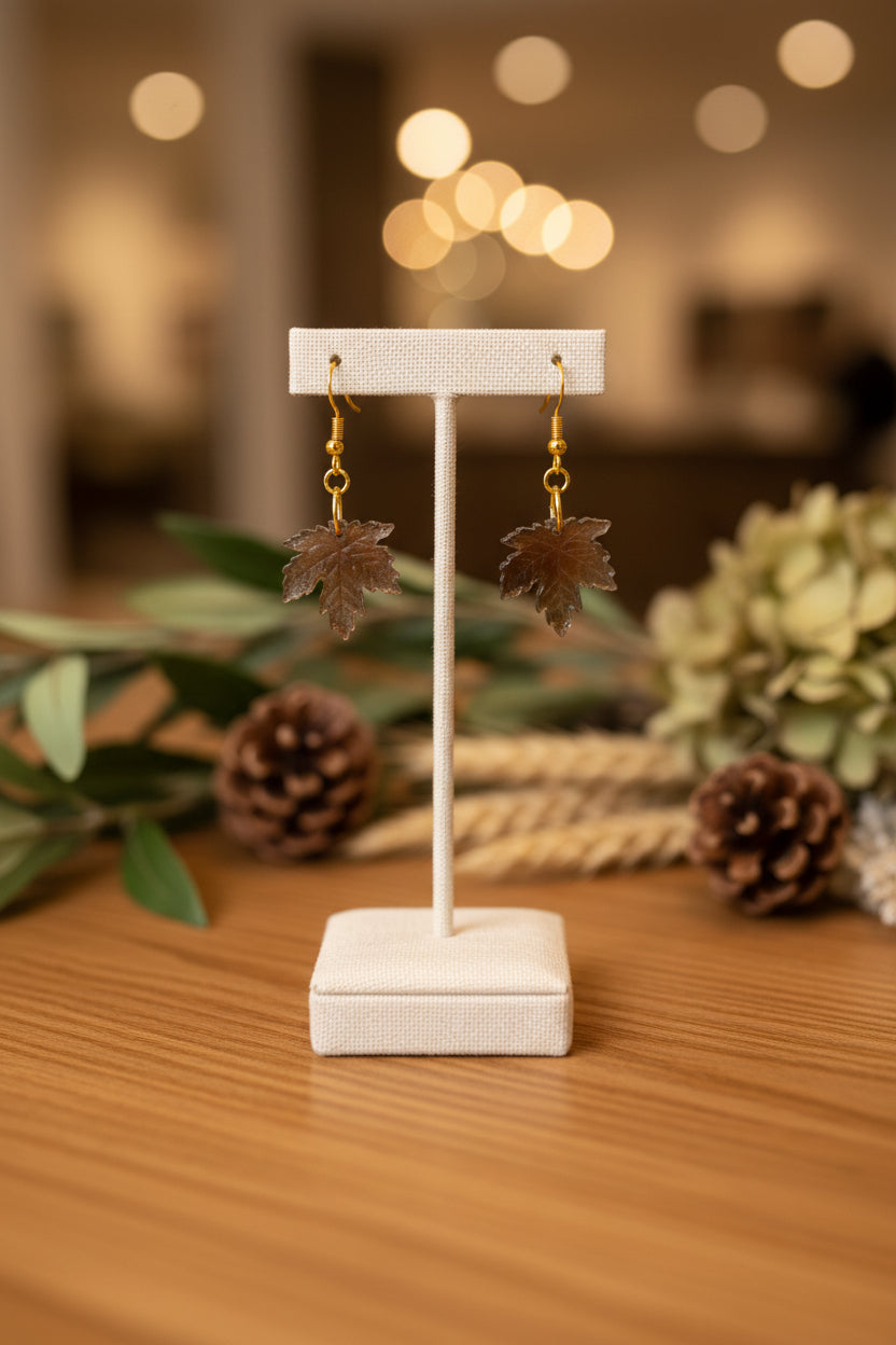 A pair of dangling earrings with brown leaf-shaped charms and gold hooks, displayed on a clear stand against a light gray background.