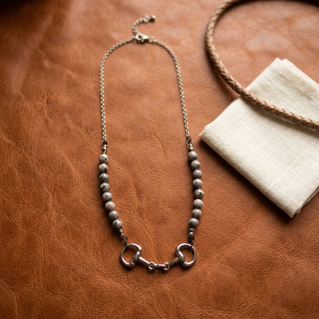 A silver horsebit necklace with gray marbled beads, displayed on a black velvet jewelry bust against a white background.