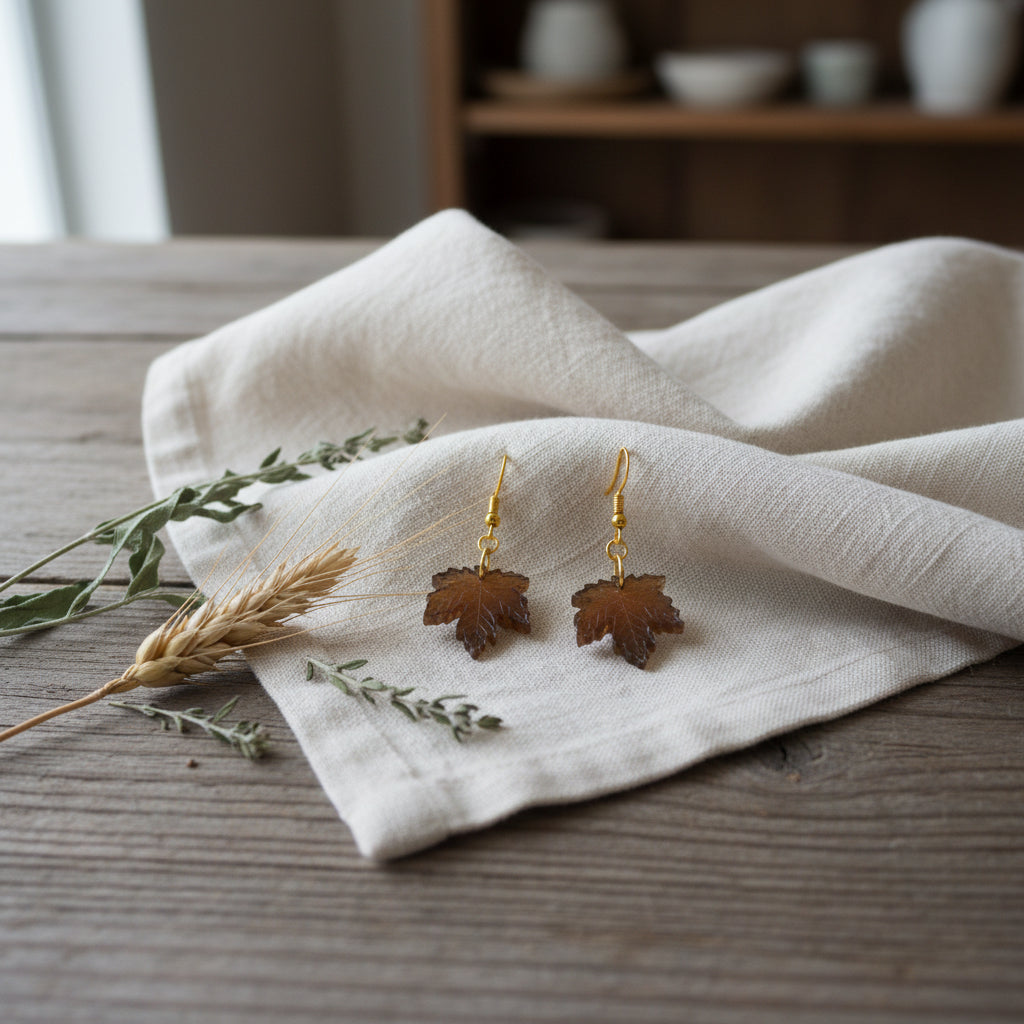 A pair of dangling earrings with small leaf-shaped charms in a warm brown tone, attached to gold-tone hooks, displayed against a plain light gray background.