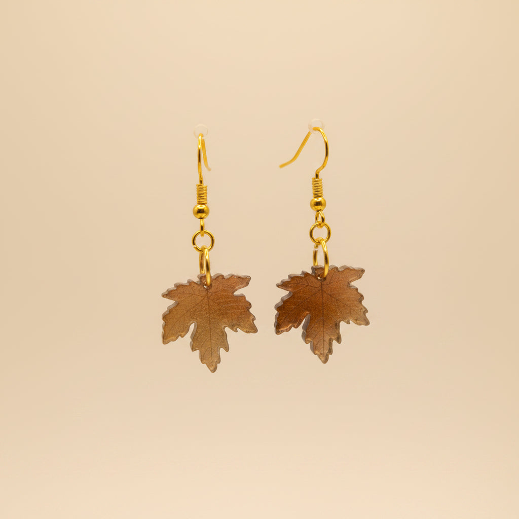 A pair of gold-tone dangle earrings with small round beads and leaf-shaped charms, displayed on a clear stand against a light gray background.