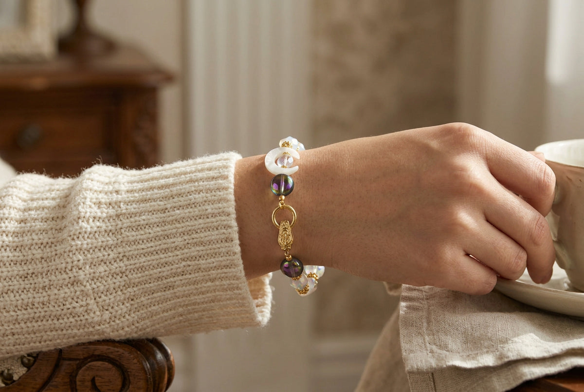 A beaded bracelet featuring white crescent moon beads, iridescent oval beads, and gold-toned spacer beads and clasp, displayed on a plain light gray background.