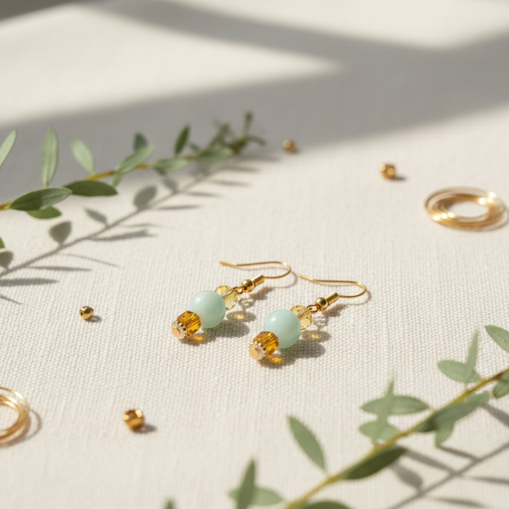 A pair of gold drop earrings featuring a light green round bead, a faceted yellow bead, and a small gold bead, displayed against a neutral gray background.