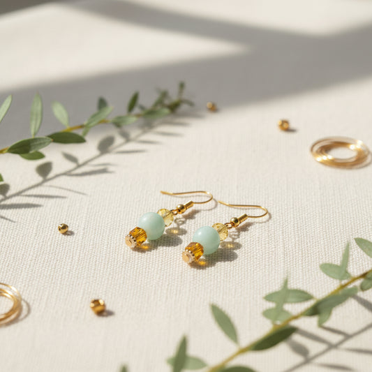 A pair of gold drop earrings featuring a light green round bead, a faceted yellow bead, and a small gold bead, displayed against a neutral gray background.