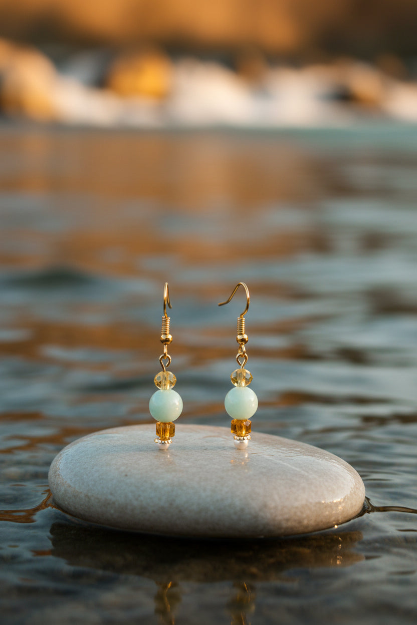 A pair of gold-tone dangle earrings with a light green bead, a small faceted gold bead, and a larger faceted amber crystal bead, displayed on a clear stand against a neutral gray background.