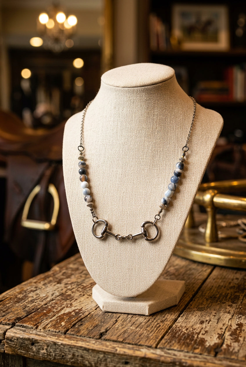 A silver-toned horse bit necklace with light blue and white marbled beads, displayed on a black velvet jewelry bust against a white background.