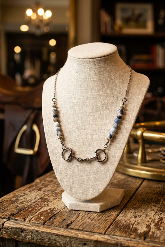 A silver-toned horse bit necklace with light blue and white marbled beads, displayed on a black velvet jewelry bust against a white background.