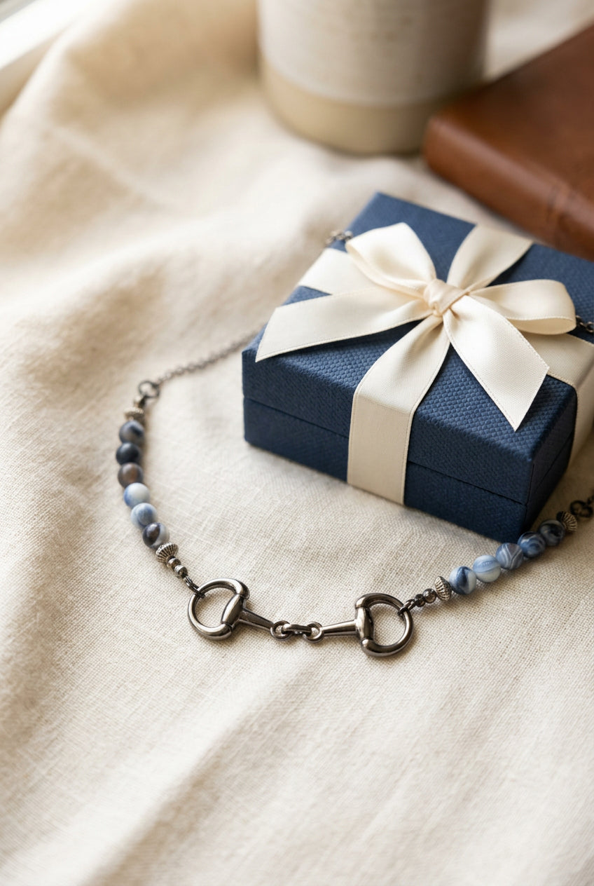 A silver-toned horse bit necklace with light blue and white marbled beads, displayed on a black velvet jewelry bust against a white background.