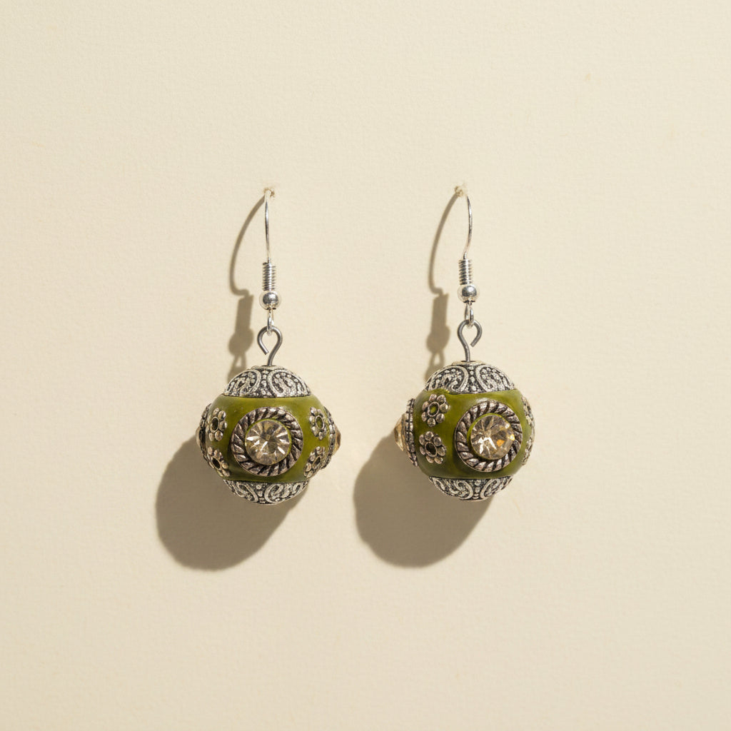 A pair of handmade dangle earrings with olive green enamel, silver filigree, and small cream-colored stones, hanging from silver fishhook wires on a clear acrylic display stand.