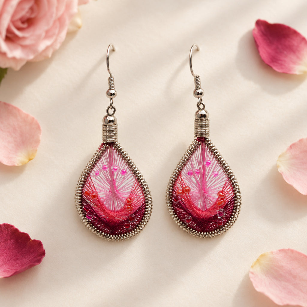 A pair of handmade teardrop dangle earrings with pink and red floral resin inlays, wrapped in silver-toned wire, hanging from silver fishhook ear wires against a light gray background.