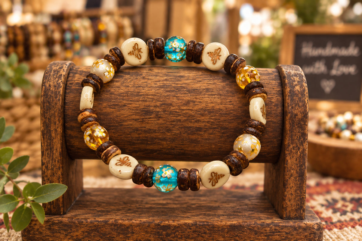 A beaded stretch bracelet featuring white beads with brown floral engravings, dark brown wooden beads, amber glass beads with gold leaf, and two blue marbled glass beads with gold flecks, displayed on a neutral gray background.