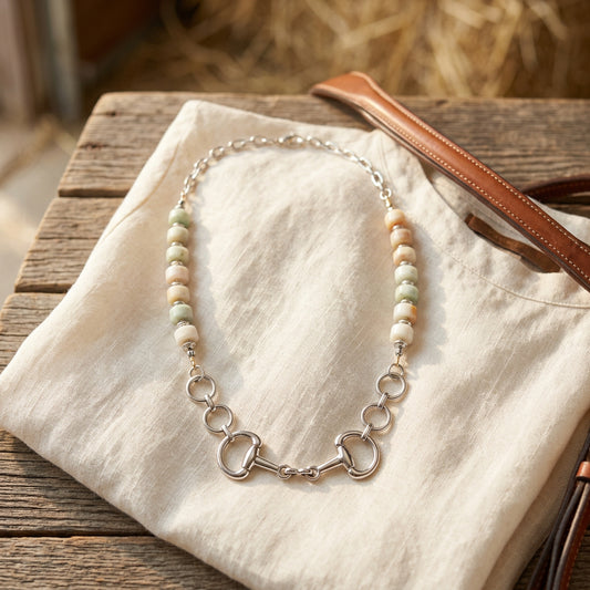 A silver horse bit necklace with white and peach-colored beads, displayed on a black velvet jewelry bust against a white background.