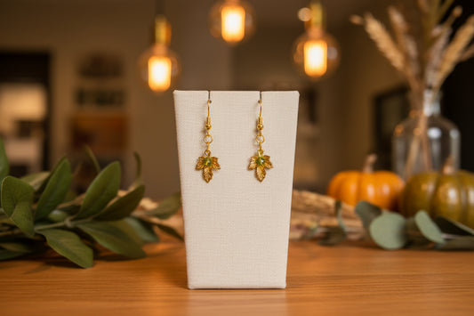 A pair of gold leaf dangle earrings displayed on a clear acrylic stand against a neutral gray background.