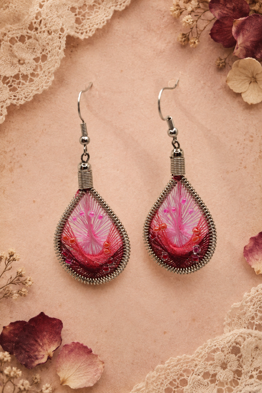 A pair of teardrop-shaped dangle earrings with silver-toned wire frames and hooks, featuring pink and red feather-like inlay with small orange accents, displayed against a light gray background.