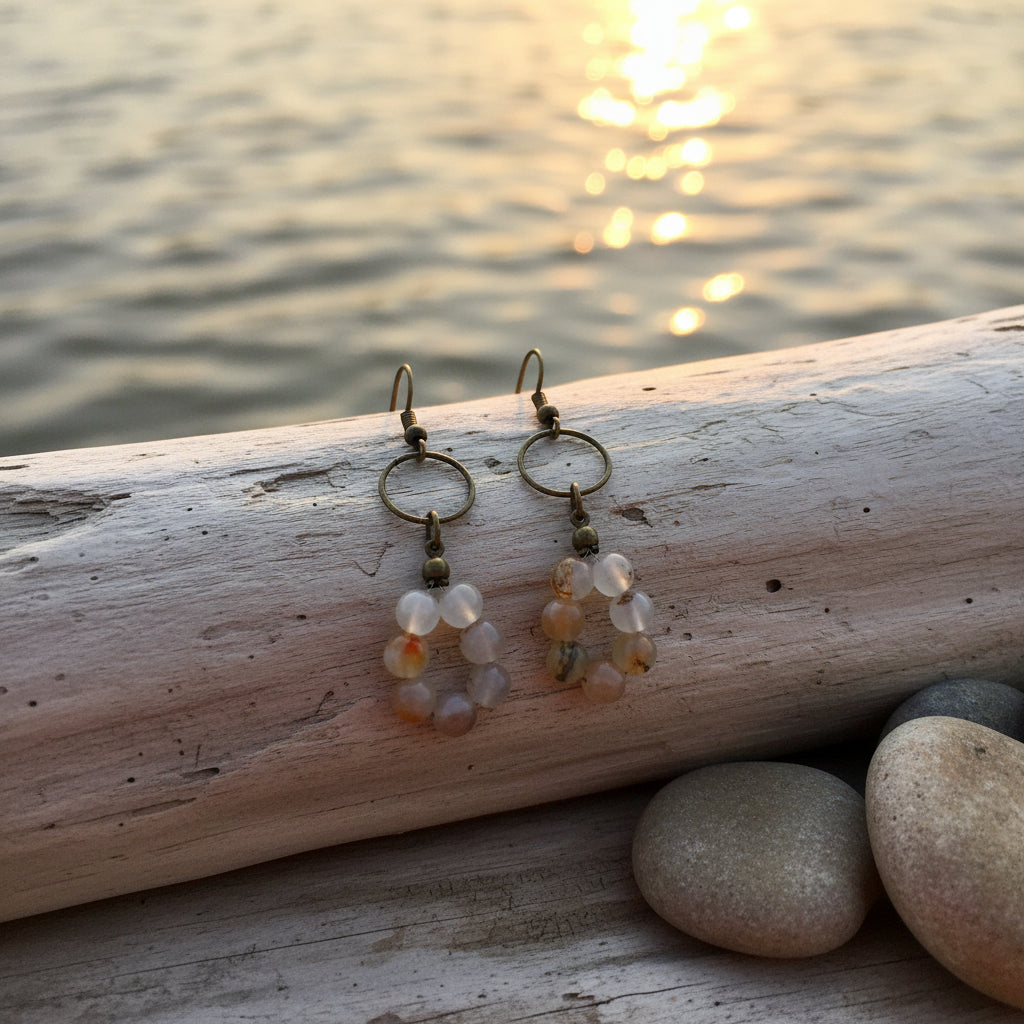 A pair of handmade dangle earrings featuring circular metal hoops and clusters of small agate beads in earthy tones of orange, brown, and cream, displayed on a clear acrylic stand against a neutral light gray background.