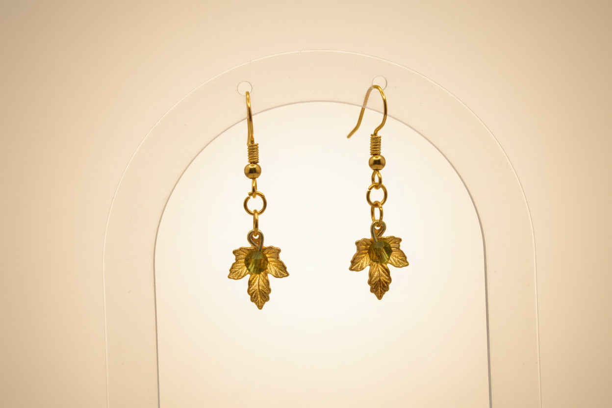 A pair of gold leaf dangle earrings with small green bead accents, displayed on a clear plastic stand against a light gray background.