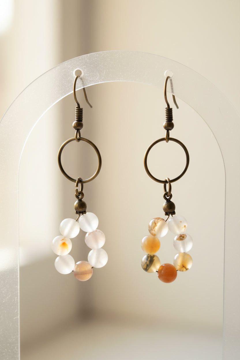 A pair of handmade dangle earrings with dark metal rings and small translucent beads in white, clear, and orange, displayed on a white stand against a white background.