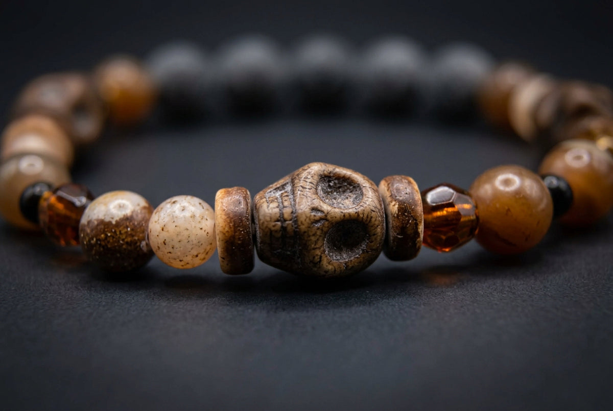 A beaded necklace featuring carved dark brown skull beads, polished amber-brown round beads, translucent light beige beads, and small black and yellow accent beads, displayed on a neutral light gray background.