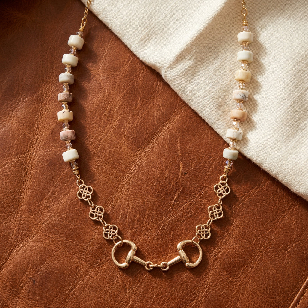 A gold-tone horsebit necklace with white and pinkish-beige faceted beads, displayed on a black velvet jewelry bust against a white background.