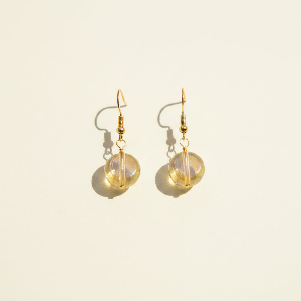 A pair of gold-tone dangle earrings with round, iridescent opal-like beads, displayed on a clear acrylic stand against a neutral gray background.