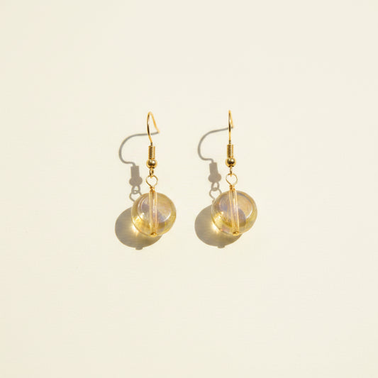 A pair of gold-tone dangle earrings with round, iridescent opal-like beads, displayed on a clear acrylic stand against a neutral gray background.